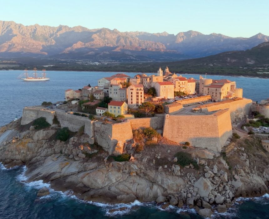 Une ville fortifiée se trouve sur une péninsule rocheuse entourée par la mer, avec des murs de pierre, des bâtiments groupés et des montagnes en arrière-plan - une vue pittoresque le long de la route cycliste GT20 Corse Nord. Un grand voilier est visible sur l'eau près de la ville. - Ekilib