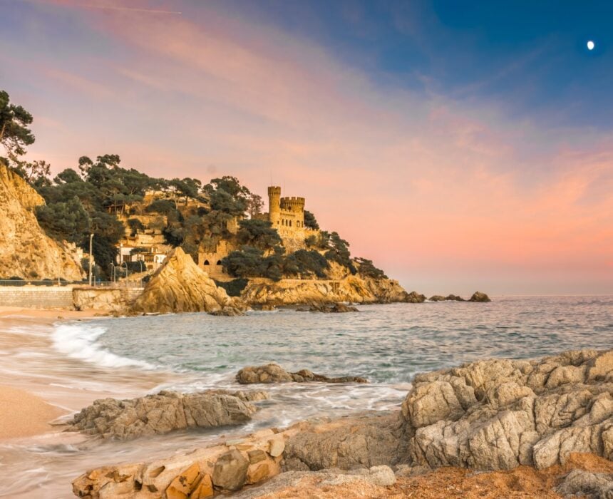 Une scène côtière au coucher du soleil avec des vagues frappant le rivage rocheux, une plage de sable et un château en pierre entouré d'arbres sur une falaise. Le ciel rose et bleu, la lune visible et les Pyrénées méditerranéennes lointaines évoquent un sentiment d'aventure pour tout amateur de vélo. - Ekilib