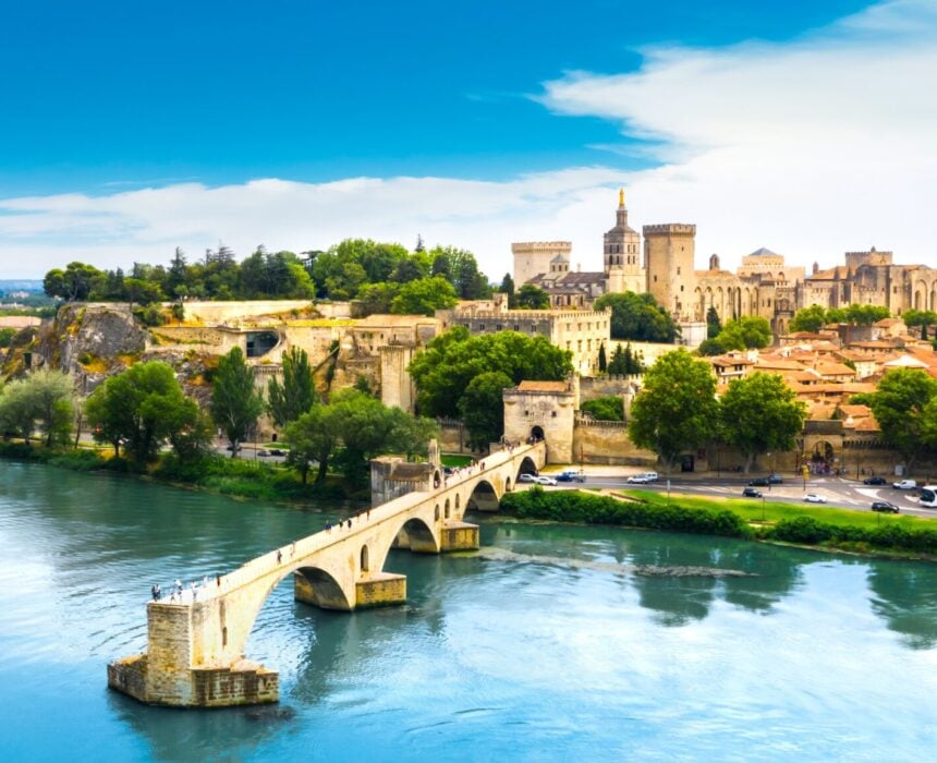 Un pont de pierre historique traverse une large rivière, menant à une ville aux bâtiments bronzés, aux arbres verts et à un grand palais médiéval - parfait pour une exploration en vélo de provence - sous un ciel bleu avec des nuages épars. - Ekilib