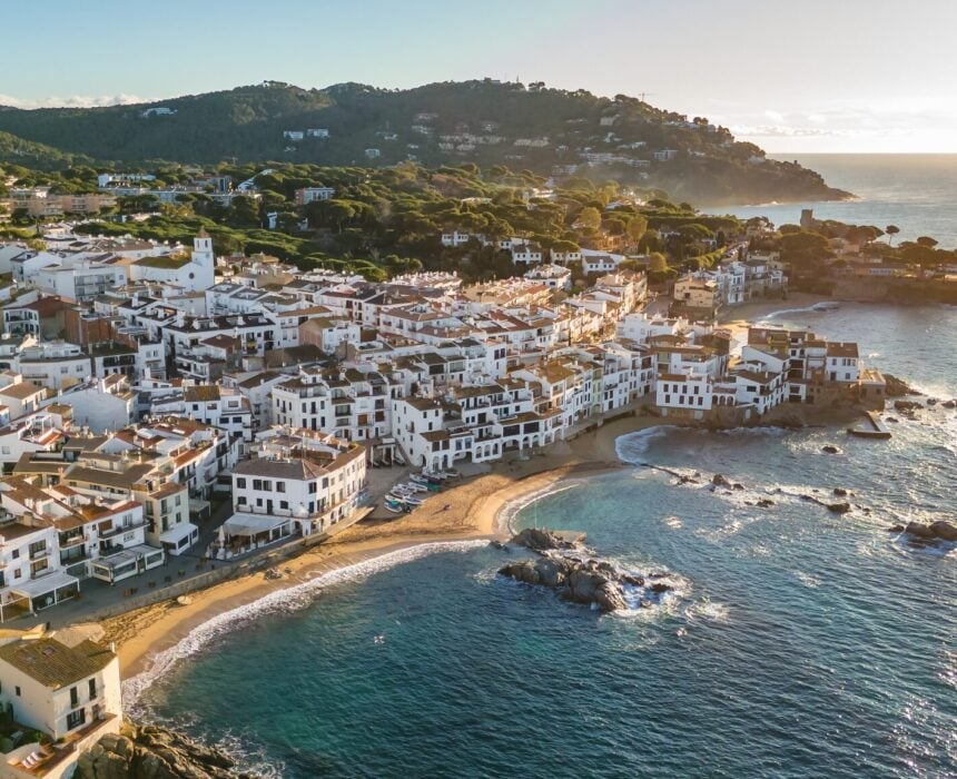 Vue aérienne d'une ville côtière de Gerone Emporda avec des bâtiments blancs regroupés le long d'une plage de sable et d'un rivage rocheux, bordés par une mer bleue claire et des collines vertes sous un ciel clair au coucher du soleil - parfait pour les amateurs de vélo. - Ekilib