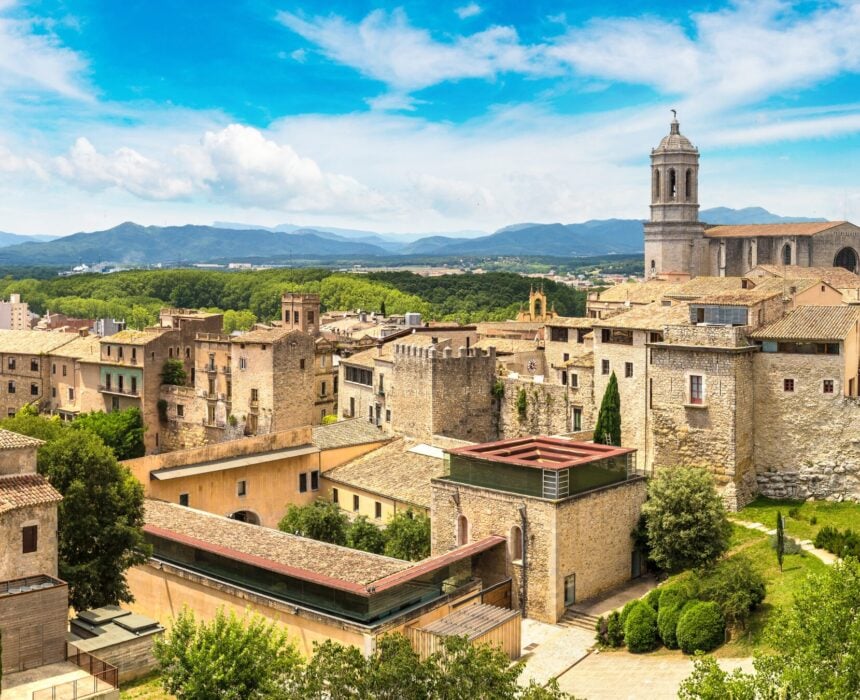 Vue panoramique d'une ville historique européenne en Catalogne avec des bâtiments en pierre, une grande cathédrale et son clocher, de la verdure, des montagnes lointaines et des cyclistes profitant de la scène sur un vélo électrique sous un ciel bleu parsemé de nuages. - Ekilib