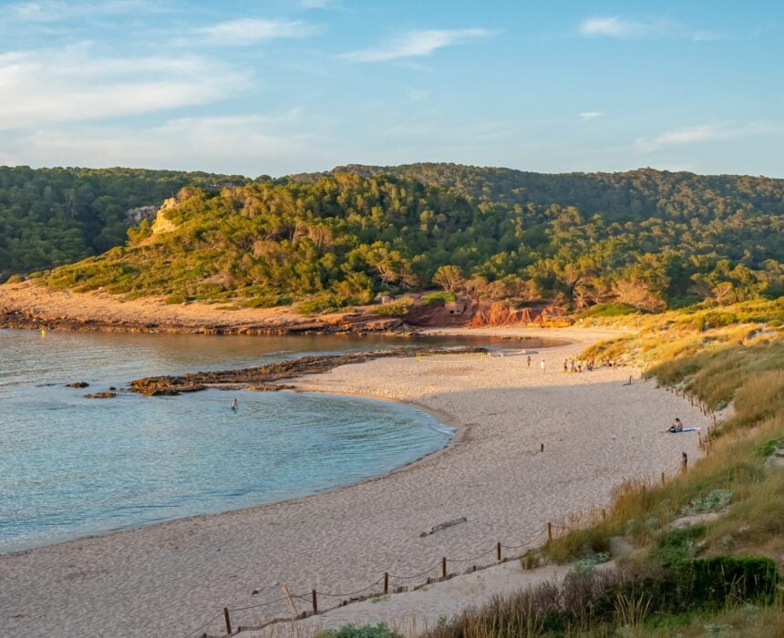 Une plage de sable s'étire le long d'une baie calme aux eaux bleues et limpides, bordée de dunes herbeuses et de collines verdoyantes couvertes d'arbres, sous un ciel partiellement nuageux au coucher du soleil. Quelques personnes et un vélo mineur se reposent tranquillement sur le rivage. - Ekilib
