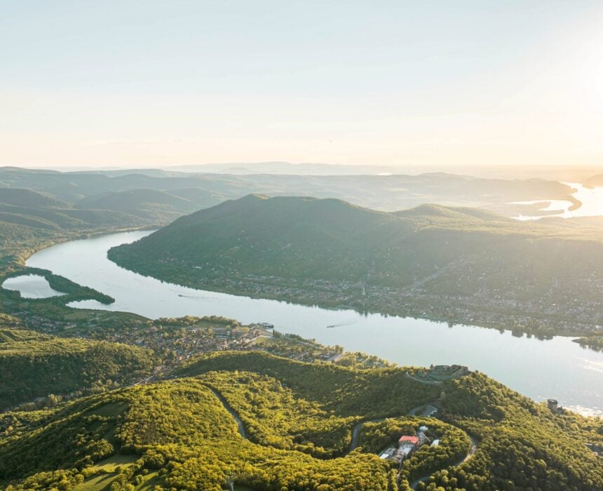 Une large rivière serpente à travers un paysage vert et vallonné sous un ciel clair. Le soleil est bas sur l'horizon, éclairant l'eau et les collines près de Velo Vienne, une route panoramique entre Bratislava et Budapest, parsemée de bâtiments épars. - Ekilib