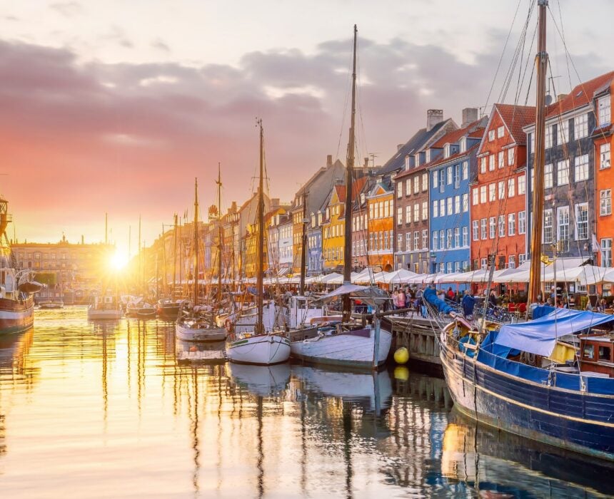 Bateaux amarrés le long d'un canal à Seeland Sud, avec des bâtiments colorés et des cafés en plein air le long du front de mer au coucher du soleil. Le soleil jette une lueur chaude, se reflétant dans l'eau - un endroit idéal pour faire une pause lors d'une balade en vélo. - Ekilib