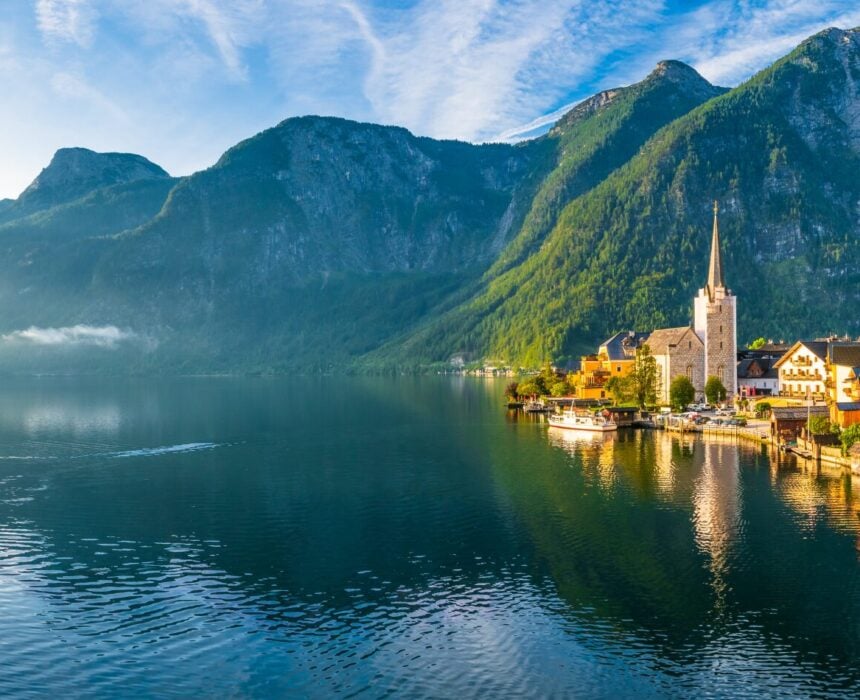 Un village lacustre le long de la Route des lacs autrichiens, avec des bâtiments colorés, une église à forte pente et des montagnes en arrière-plan, sous un ciel partiellement nuageux. L'eau calme reflète le charme du paysage. - Ekilib