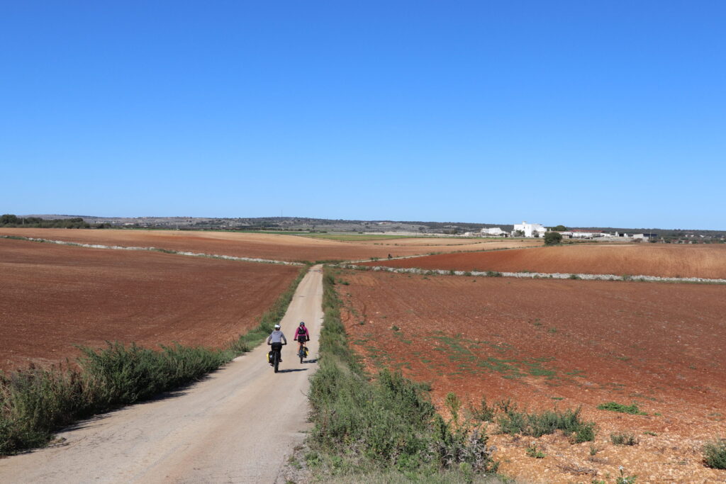 Deux personnes à vélo sur une route de campagne étroite traversant de vastes champs labourés de couleur brun-rouge sous un ciel bleu clair dans la région des Pouilles à vélo, avec des bâtiments lointains visibles à l'horizon. - Ekilib