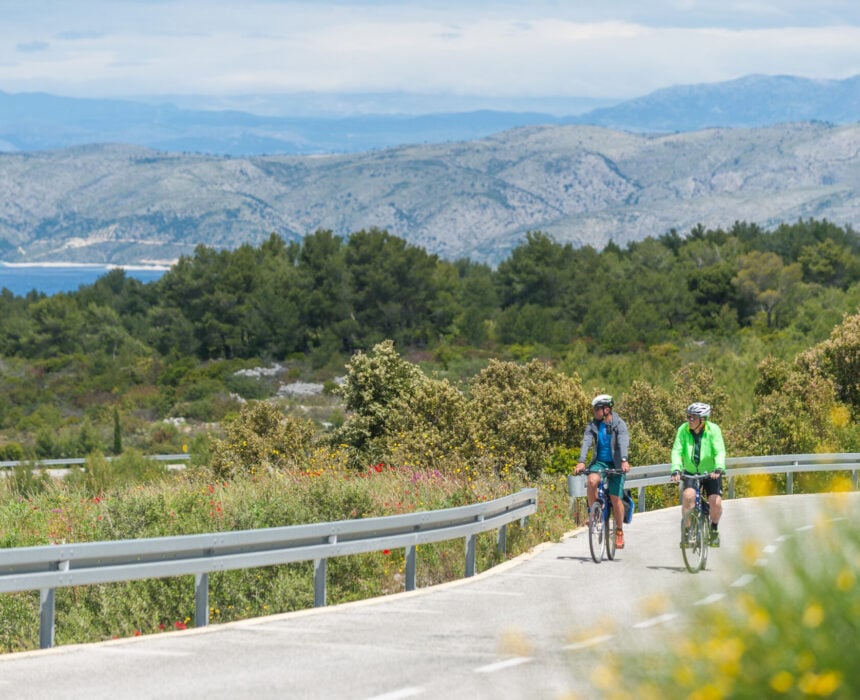 Deux personnes portant des casques et des vêtements de sport conduisent des vélos sur une route sinueuse entourée de verdure, avec des collines, des arbres et de l'eau en arrière-plan, évoquant la beauté des paysages de la Croatie sous un ciel partiellement nuageux. - Ekilib