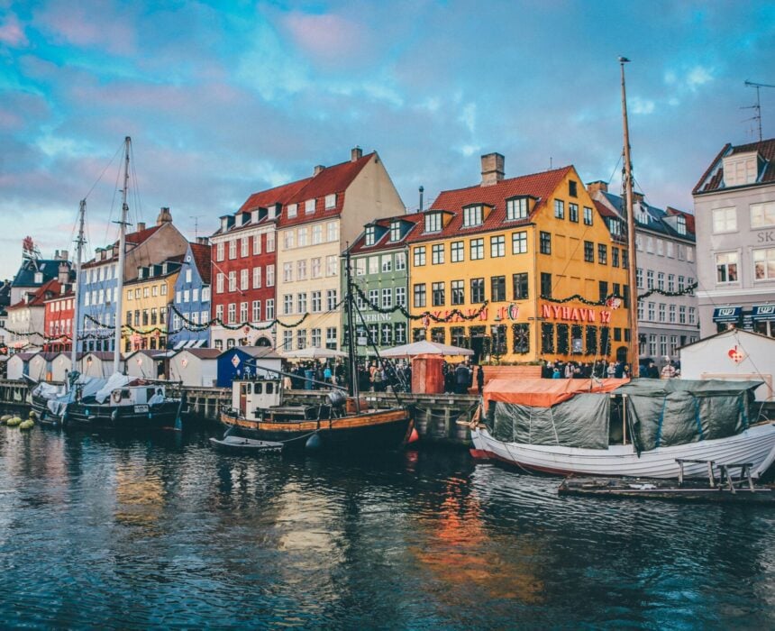 Des bâtiments colorés bordent une rue au bord de l'eau dans le quartier de Nyhavn à Copenhague. Des bateaux accostent le long du canal, tandis que des gens se promènent ou se détendent dans des cafés en plein air - une halte invitante pour ceux qui explorent Seeland à vélo sous un ciel partiellement nuageux. - Ekilib