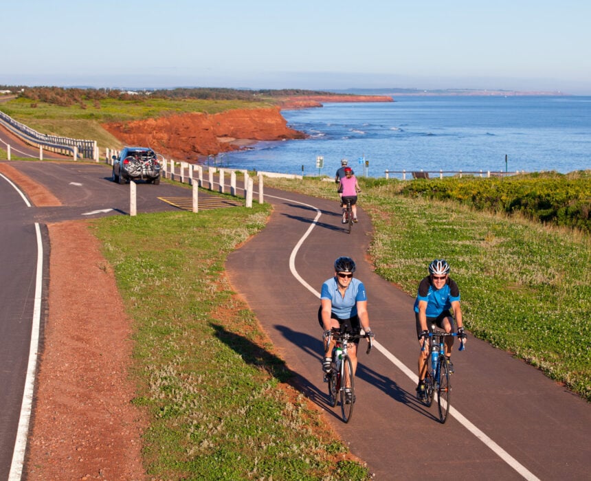 Trois cyclistes roulent sur un chemin pavé le long de l'île du Prince Édouard, avec des zones herbeuses et des falaises rouges au bord de l'océan. Une voiture garée près de la route sous un ciel clair, parfait pour une journée de vélo sur la côte. - Ekilib