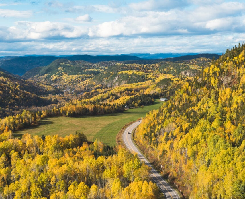 Une route sinueuse traverse un paysage de collines couvertes d'arbres d'automne jaunes et verts sous un ciel partiellement nuageux, avec des montagnes en arrière-plan - évoquant la beauté pittoresque du fjord Saguenay lors d'un voyage à vélo mémorable. - Ekilib