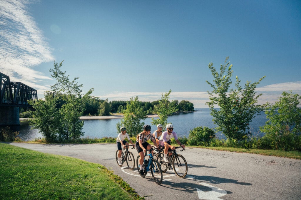 Trois personnes roulent à vélo sur un chemin pavé près d'un plan d'eau, avec des arbres verts et un pont en arrière-plan sous un ciel bleu clair - une inspiration parfaite pour votre prochain voyage avec une agence voyage vélo. - Ekilib