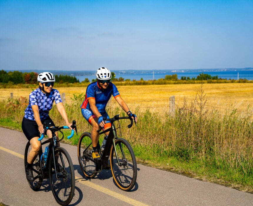 Deux personnes portant des casques et des équipements cyclistes roulent à vélo sur un chemin pavé à côté d'un champ, avec des arbres et le Lac Saint Jean visibles en arrière-plan sous un ciel bleu clair. - Ekilib