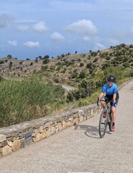 Une personne portant une chemise bleue et un casque monte à vélo sur un chemin pavé avec des murs en pierre, entouré d'un terrain vallonné - capturant l'esprit du voyage à vélo sous un ciel partiellement nuageux. - Ekilib