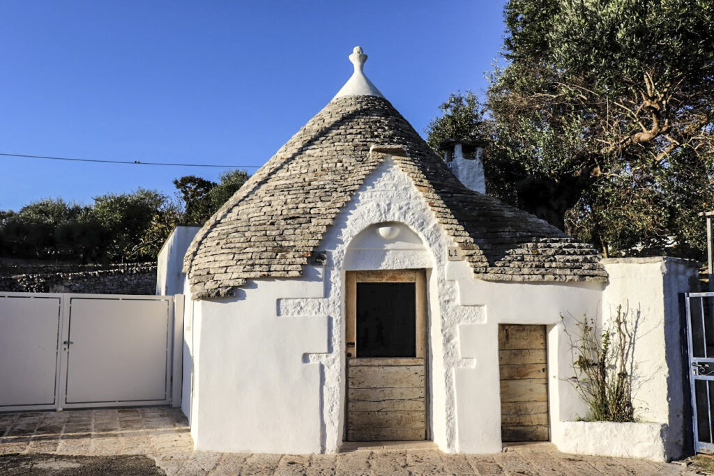Un petit bâtiment en pierre blanche avec un toit conique en pierre grise, une porte en bois et une petite porte adjacente - typique des routes de pouilles à vélo - entouré d'un muret et d'un portail, avec des arbres et un ciel bleu clair en arrière-plan. - Ekilib