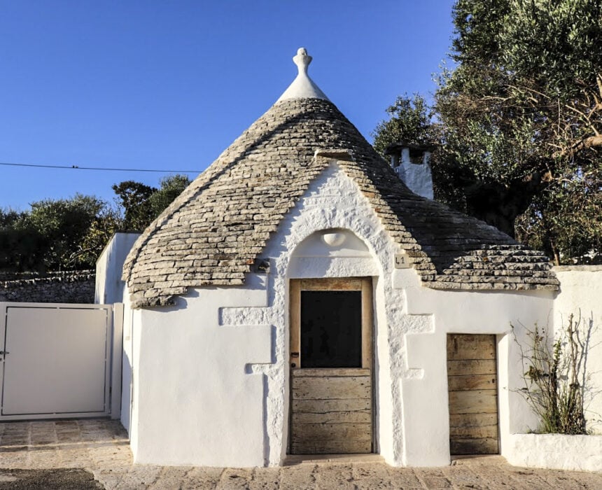 Un petit bâtiment en pierre blanche avec un toit conique en pierre grise, une porte en bois et une petite porte adjacente - typique des routes de pouilles à vélo - entouré d'un muret et d'un portail, avec des arbres et un ciel bleu clair en arrière-plan. - Ekilib
