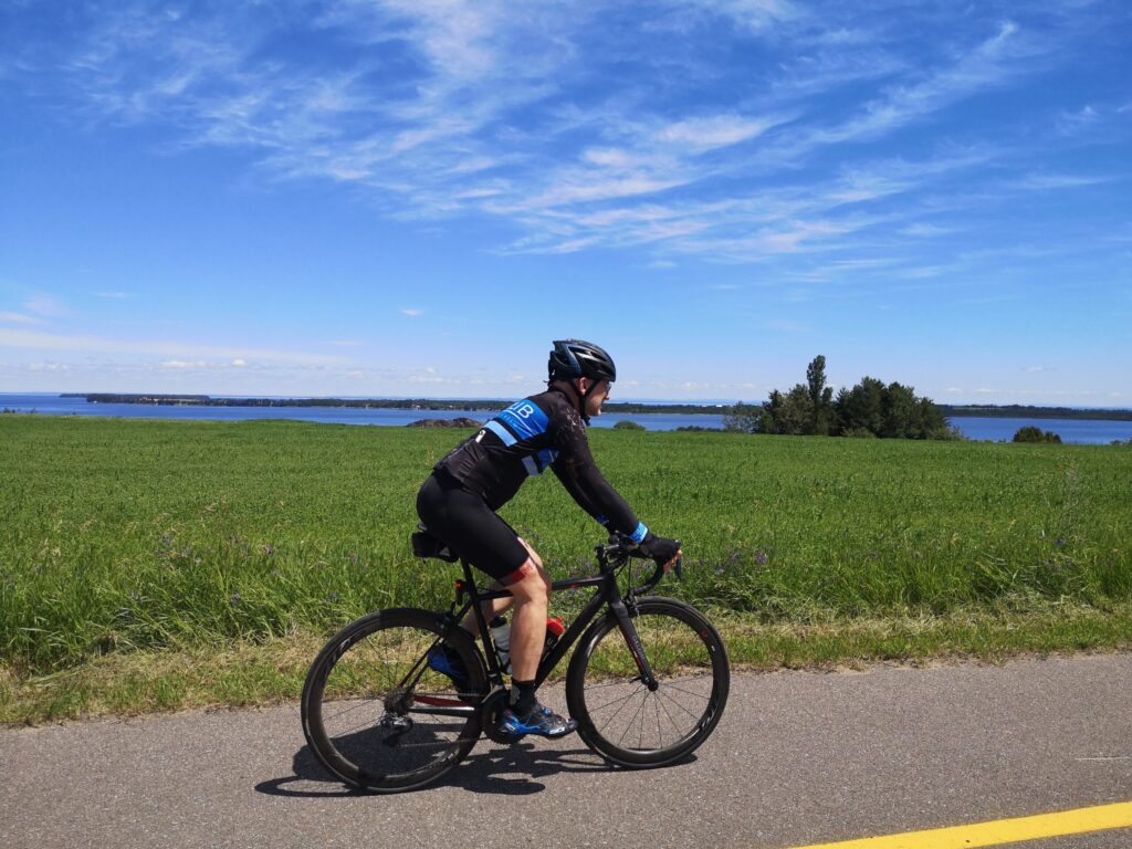 Un cycliste portant un casque et une tenue noire roule sur un vélo de route sur un chemin pavé à côté d'un champ herbeux, capturant l'esprit du Tour Lac Saint Jean Velo, avec un plan d'eau et un ciel bleu avec des nuages en arrière-plan. - Ekilib
