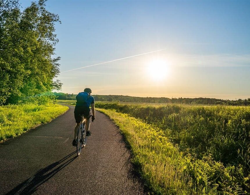 Une personne roule à vélo sur un chemin pavé dans un paysage verdoyant, avec des arbres sur la gauche et des champs ouverts sur la droite. Le soleil est bas dans le ciel, projetant de longues ombres - peut-être quelque part entre Montréal et Sherbrooke. - Ekilib