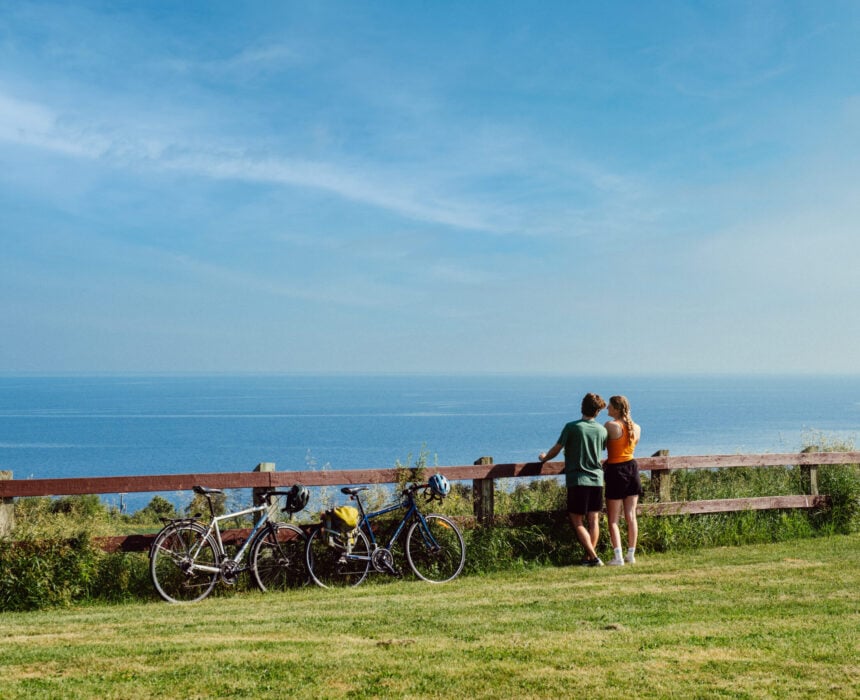 Deux personnes se tiennent près d'une barrière en bois surplombant la mer au Lac Saint Jean, avec deux vélos garés à proximité sur l'herbe sous un ciel bleu clair - parfait pour une aventure en vélo. - Ekilib