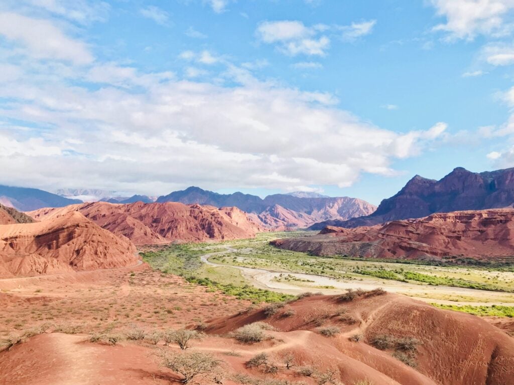 Une large vallée avec des collines rocheuses rouges et une végétation clairsemée, une rivière qui serpente à travers la plaine verdoyante, et des montagnes lointaines sous un ciel bleu - parfait pour un voyage à vélo dans les paysages à couper le souffle de l'Argentine. - Ekilib