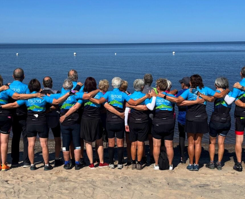 Un groupe de personnes portant des maillots cyclistes bleus assortis, participants au Tour Lac Saint Jean Vélo, se tiennent bras dessus bras dessous sur une plage de sable, regardant la mer calme sous un ciel bleu clair. - Ekilib