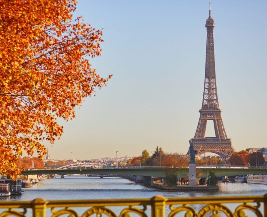 La Tour Eiffel se dresse au bord de la Seine sous un ciel clair, entourée d'arbres d'automne aux feuilles orangées. Un vélo de couleur champagne passe, tandis que des immeubles et un pont apparaissent à l'arrière-plan. - Ekilib