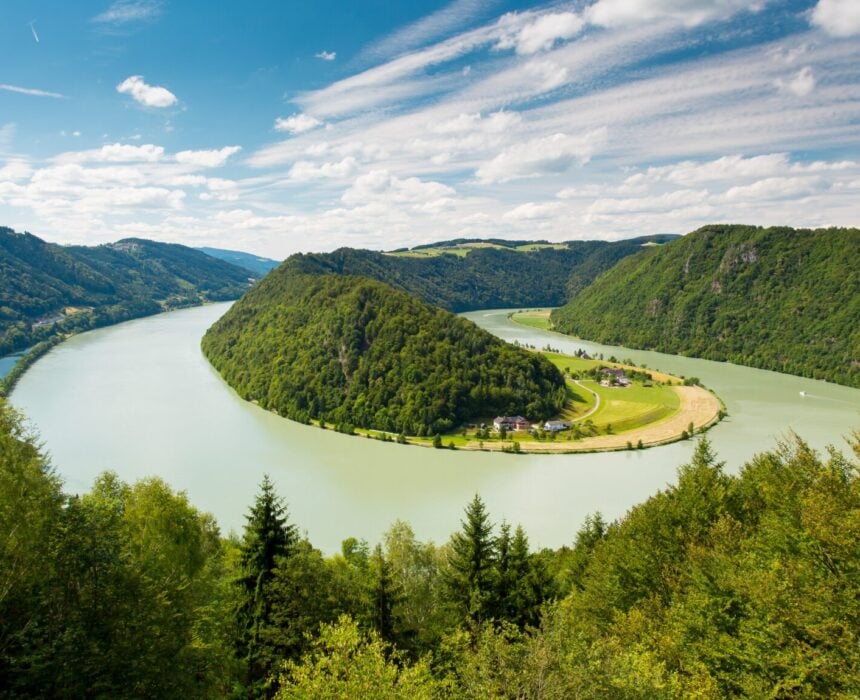 Le Danube s'enroule autour d'une péninsule vallonnée et densément boisée, avec des prairies verdoyantes à sa base, parfaite pour une exploration en vélo ou en bateau, entourée de collines couvertes d'arbres sous un ciel bleu parsemé de nuages épars. - Ekilib
