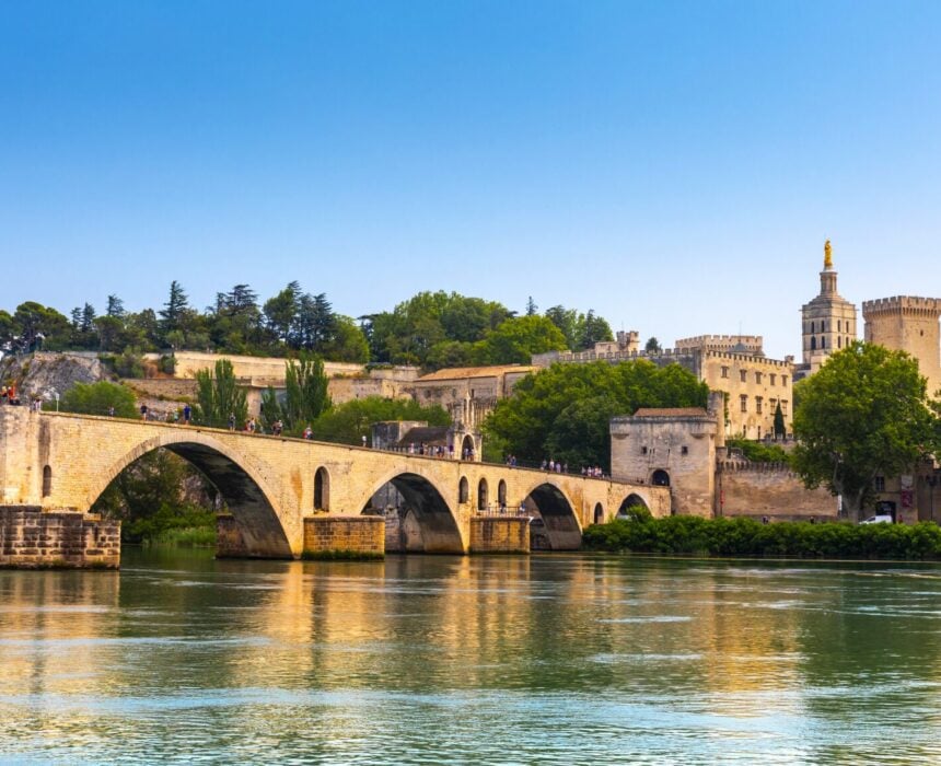 Un pont en arc de pierre traverse une rivière calme en Provence, avec une ville historique fortifiée et une tour coiffée d'or au-delà. Des arbres bordent les rives, tandis qu'un vélo et un bateau ajoutent du charme sous le ciel bleu clair. - Ekilib