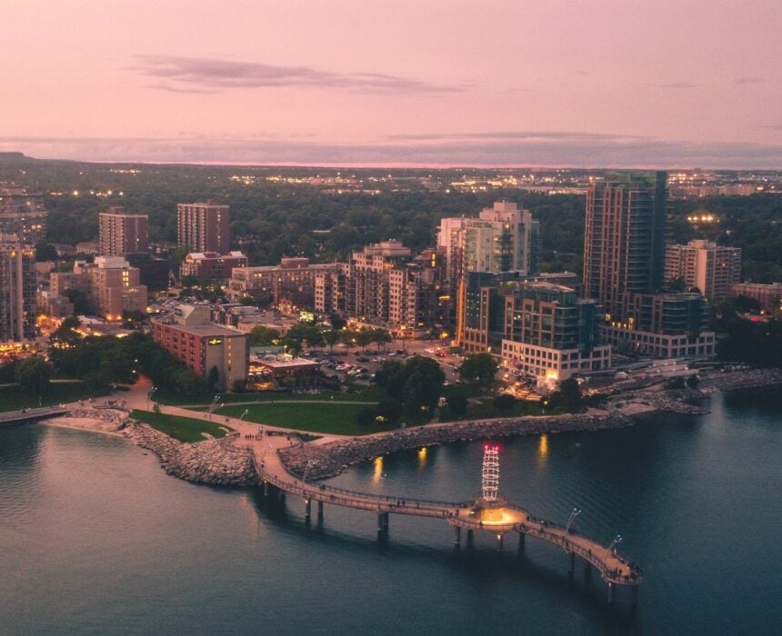 Vue aérienne d'une ville au bord de l'eau au crépuscule, avec des bâtiments, des arbres, la route du Niagara Velo serpentant près d'une jetée incurvée s'avançant dans l'eau calme, et des lampadaires commençant à éclairer la scène. - Ekilib
