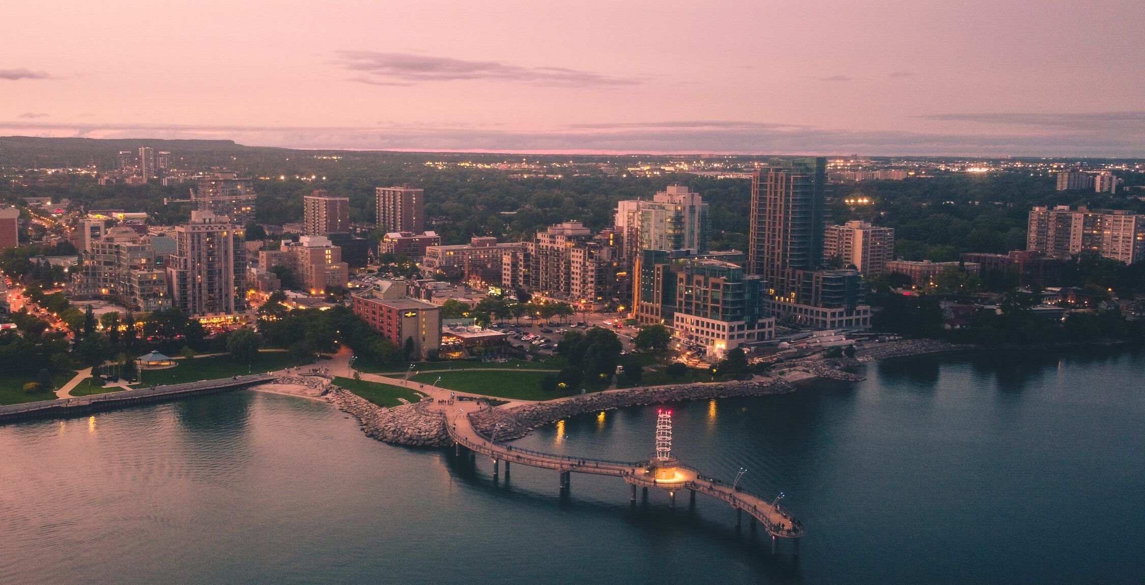 Vue aérienne d'une ville au bord de l'eau au crépuscule, avec des bâtiments, des arbres, la route du Niagara Velo serpentant près d'une jetée incurvée s'avançant dans l'eau calme, et des lampadaires commençant à éclairer la scène. - Ekilib