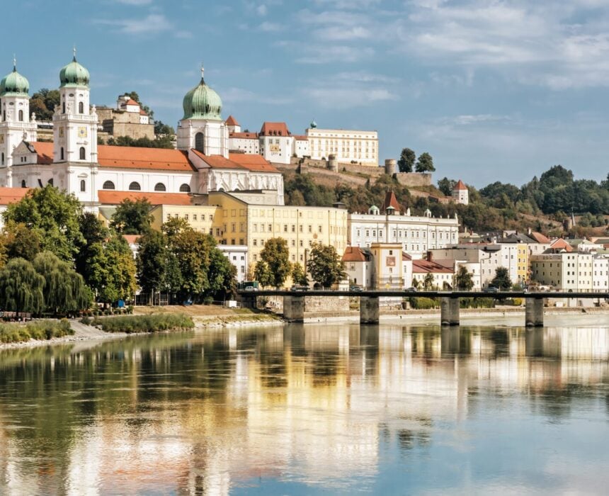 Vue d'une ville européenne en Autriche avec des bâtiments historiques, des tours d'église en forme de dôme et un pont traversant le calme fleuve Danube. Un vélo et un bateau passent, les arbres et les reflets scintillent au bord de l'eau sous un ciel partiellement nuageux. - Ekilib