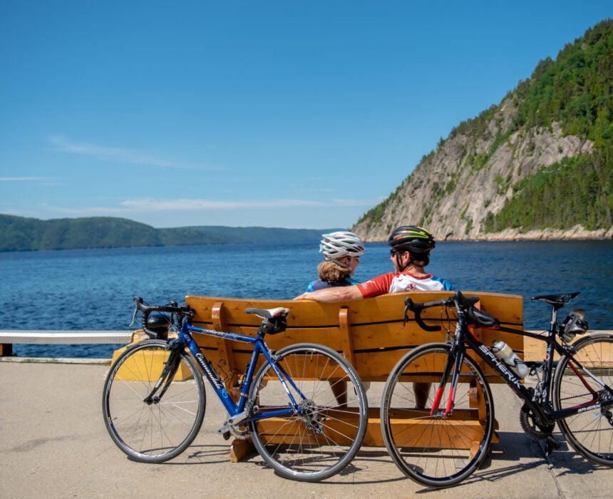 Deux personnes portant des casques de vélo sont assises sur un banc en bois face au fjord Saguenay, avec deux vélos garés à proximité. Des collines couvertes d'arbres s'élèvent au-dessus de l'eau sous un ciel bleu clair - un moment idyllique à vélo. - Ekilib