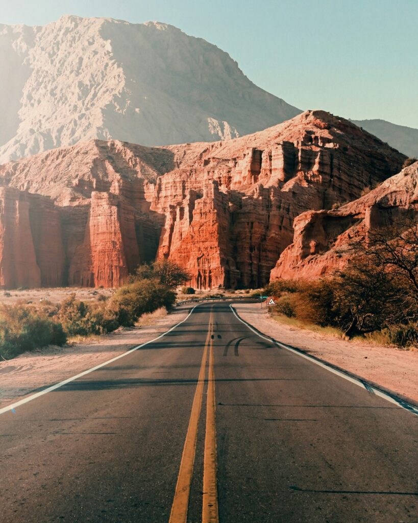 Une route pavée aux lignes jaunes s'étire vers des formations rocheuses rouges et une grande montagne à l'arrière-plan, sous un ciel clair - une scène invitante pour un voyage à vélo en Argentine, avec une végétation clairsemée qui borde la route. - Ekilib