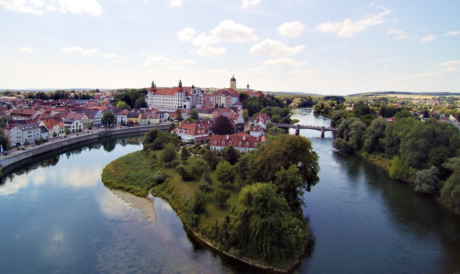 Vue d'une ville avec des bâtiments aux toits rouges le long du Danube, une île avec des arbres au premier plan et un pont traversant l'eau, sous un ciel partiellement nuageux - une scène parfaite pour les voyages en vélo-bateau. - Ekilib
