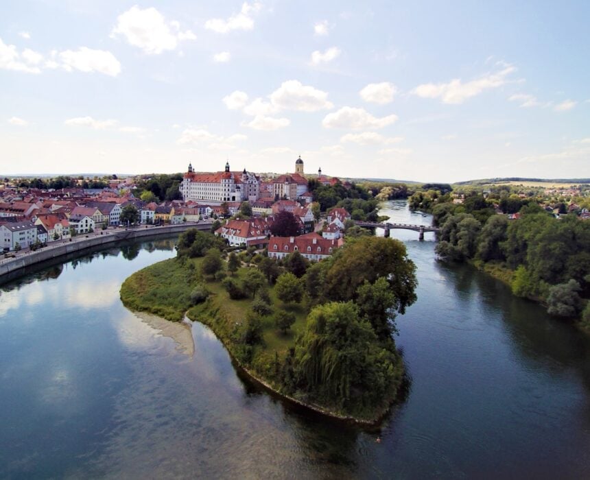 Vue d'une ville avec des bâtiments aux toits rouges le long du Danube, une île avec des arbres au premier plan et un pont traversant l'eau, sous un ciel partiellement nuageux - une scène parfaite pour les voyages en vélo-bateau. - Ekilib
