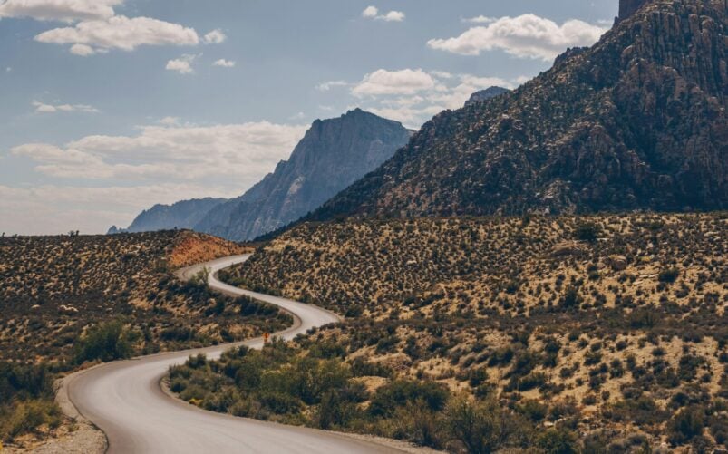 Une route sinueuse traverse un paysage désertique sec et rocailleux à la végétation clairsemée. Des montagnes s'élèvent à l'arrière-plan sous un ciel partiellement nuageux. - Ekilib
