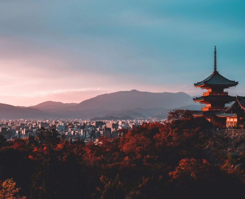 Une pagode japonaise traditionnelle est située sur une colline au feuillage d'automne rouge, surplombant un paysage urbain et le pittoresque Shiminami Kaido au coucher du soleil. Des montagnes sont visibles au loin sous un ciel partiellement nuageux. - Ekilib