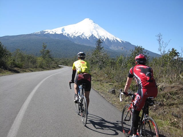 Deux cyclistes roulent sur une route pavée bordée de verdure, avec une montagne enneigée en arrière-plan, sous un ciel bleu clair. - Ekilib