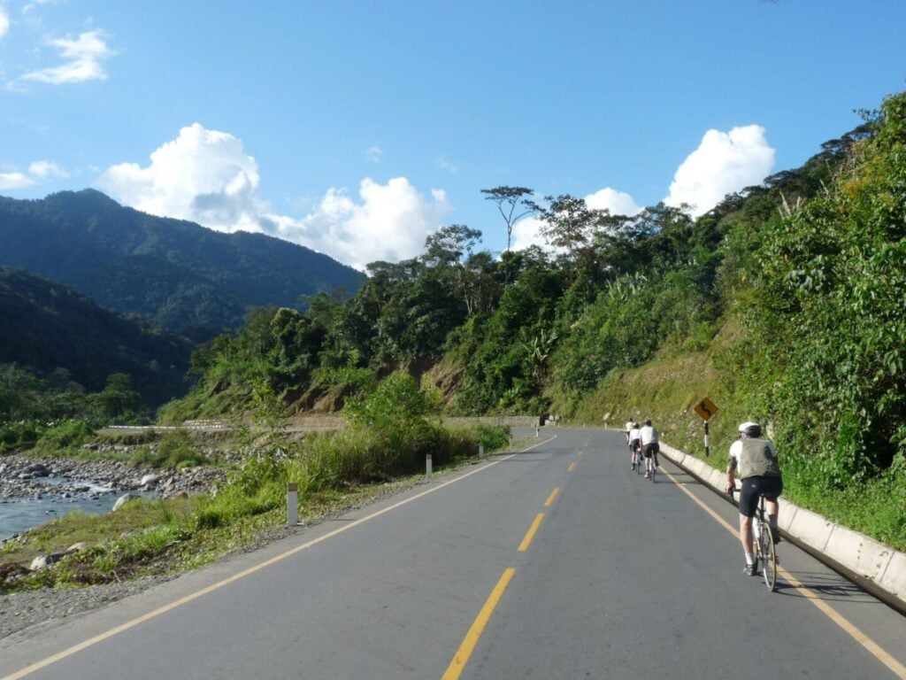 Un groupe de cyclistes roule sur une route pavée bordée d'arbres verdoyants et de montagnes, avec une rivière sur le côté gauche et un ciel partiellement nuageux au-dessus de la tête. - Ekilib