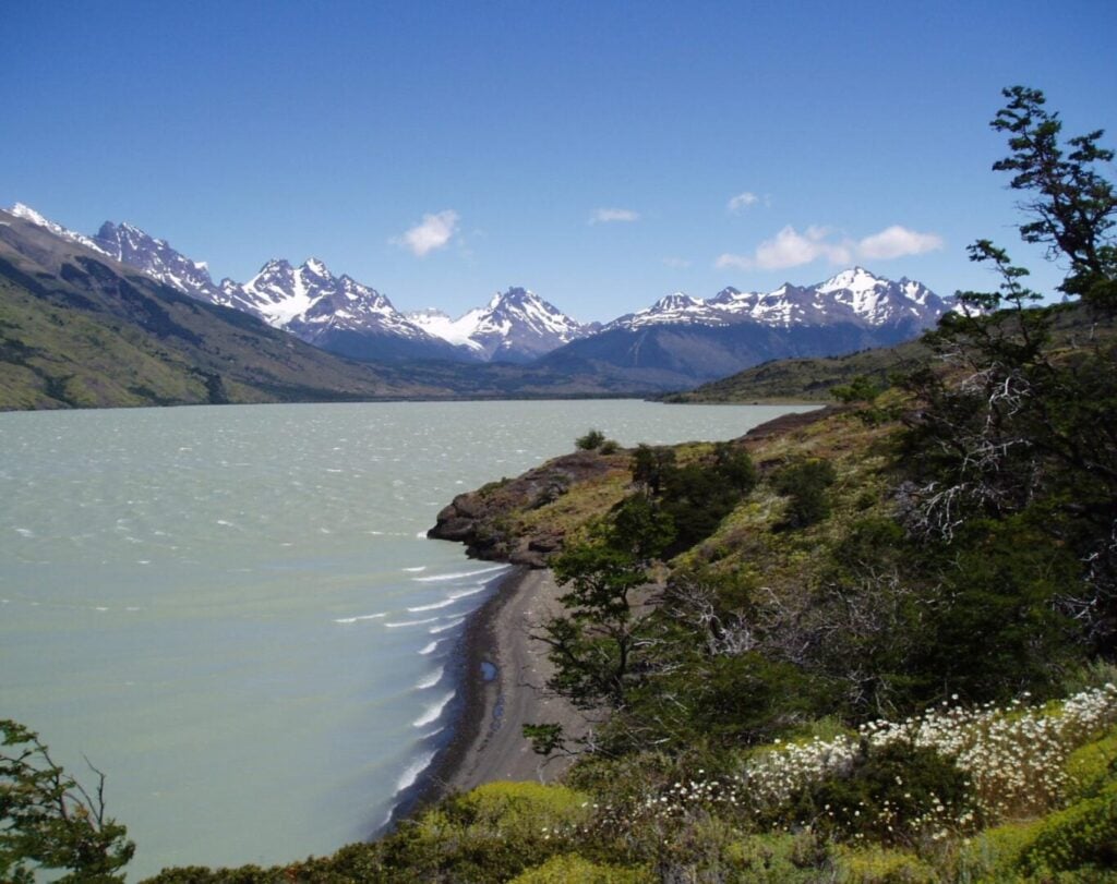 Un lac aux eaux vert clair s'enroule autour d'un rivage rocheux, avec des montagnes enneigées à l'arrière-plan et une végétation verdoyante au premier plan sous un ciel bleu. - Ekilib