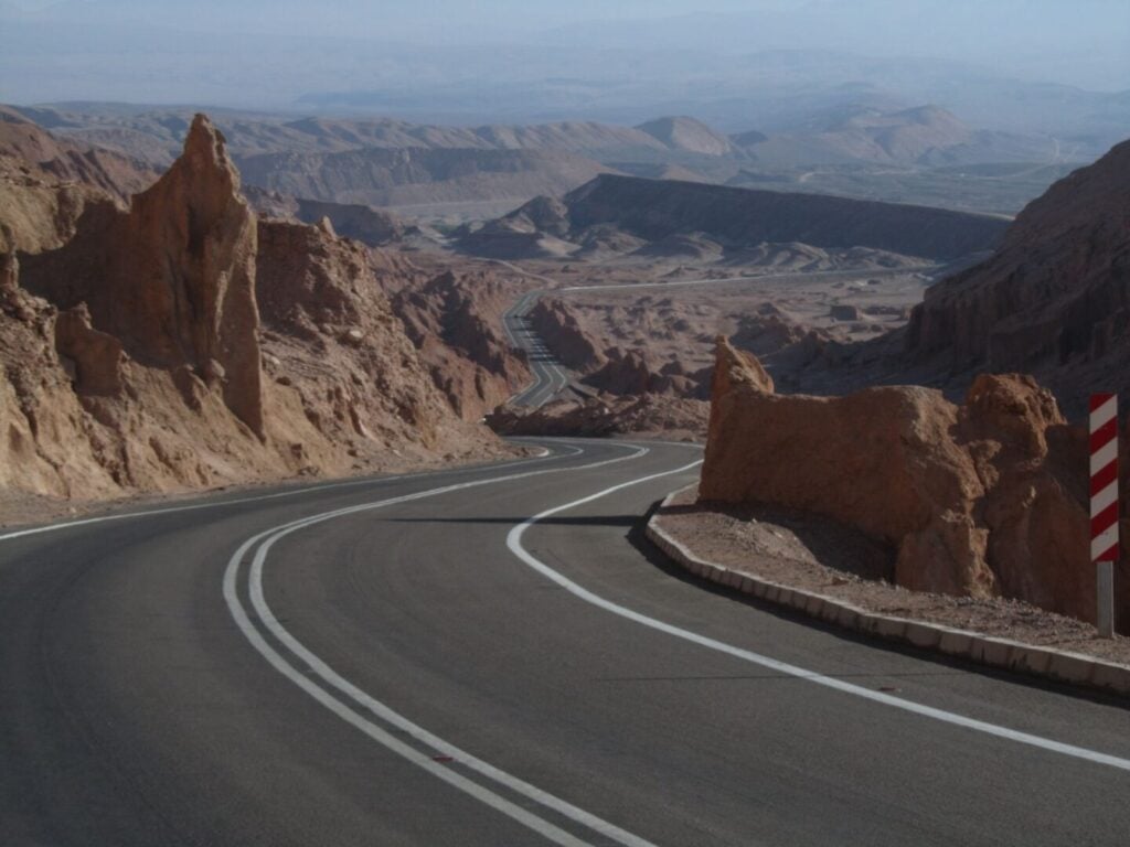 Une route sinueuse avec des lignes blanches doubles traverse un paysage rocheux et aride avec des collines érodées et des montagnes lointaines sous un ciel brumeux. Un panneau de signalisation rouge et blanc est visible sur le côté droit. - Ekilib