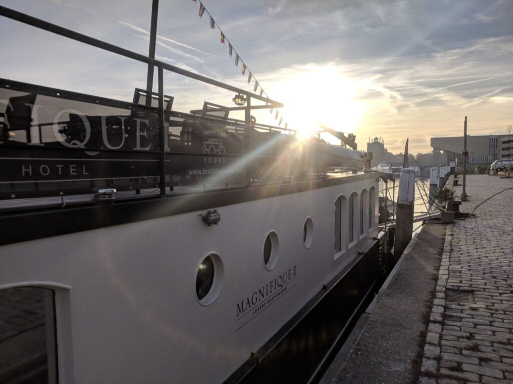 Un bateau blanc portant l'inscription « MAGNETIQUE » est amarré à un quai pavé, tôt le matin ou en fin d'après-midi, alors que le soleil est bas dans le ciel et projette de longues ombres. - Ekilib