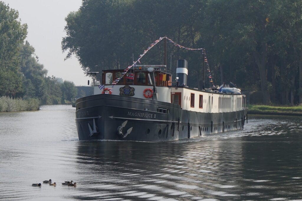 Une péniche en noir et blanc baptisée MAGNIFIQUE II avance sur un cours d'eau calme bordé d'arbres, avec quelques canards nageant au premier plan et des guirlandes festives accrochées le long du bateau. - Ekilib