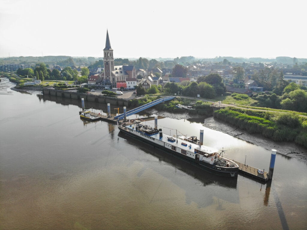Un ferry de passagers est amarré à un embarcadère sur une rivière calme ; en arrière-plan, sous un ciel brumeux, on aperçoit une petite ville, une église au clocher élancé et de la verdure. - Ekilib