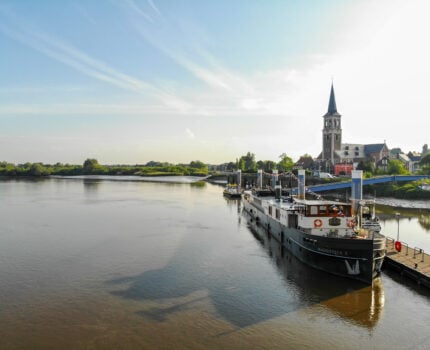 Un ferry baptisé « Rogentrans II » est amarré à un embarcadère sur une rivière calme ; en arrière-plan, sous un ciel partiellement nuageux, on aperçoit un village et une église dotée d'un haut clocher. - Ekilib