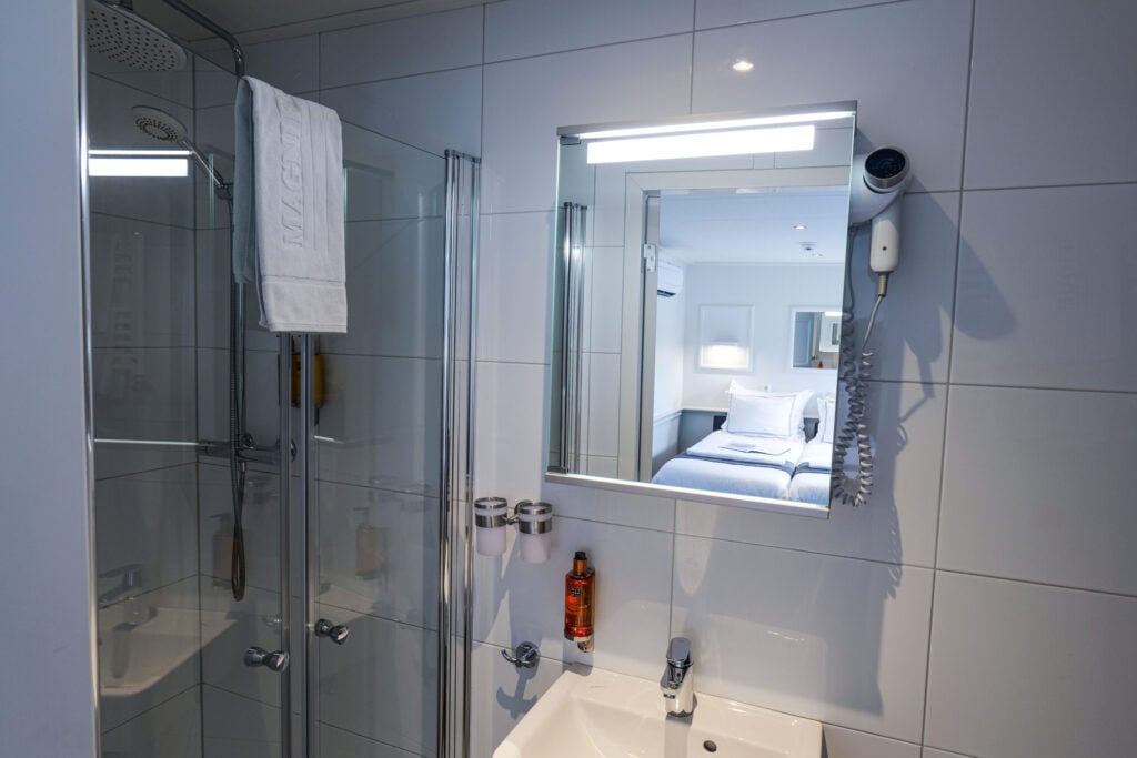 Une salle de bains moderne avec des murs carrelés de blanc, une cabine de douche en verre, une serviette accrochée à la porte de la douche, un lavabo sous un miroir, un sèche-cheveux fixé au mur, deux tasses accrochées au mur et le reflet d'un lit bien fait. - Ekilib