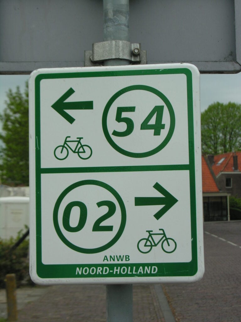 Un panneau rectangulaire représentant deux flèches vertes accompagnées de symboles de vélo : l'une pointe vers la gauche et porte le numéro 54, tandis que l'autre pointe vers la droite et porte le numéro 02. En bas du panneau figure l'inscription « ANWB NOORD-HOLLAND ». - Ekilib