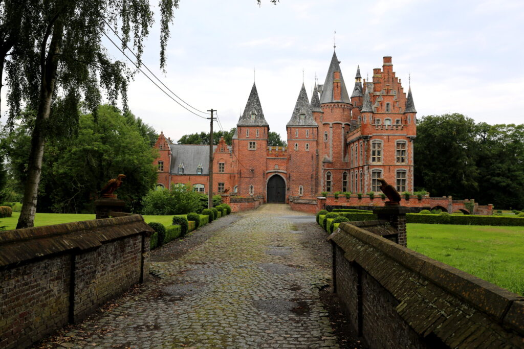 Un chemin pavé mène à un grand château en briques rouges, doté de nombreuses tours pointues et de fenêtres cintrées, entouré de pelouses verdoyantes et d'arbres. - Ekilib