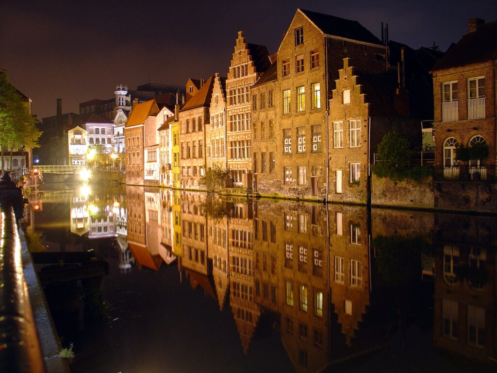 Une rangée de bâtiments historiques en briques est illuminée la nuit, leurs lumières se reflétant nettement dans un canal paisible. Le ciel est sombre et la scène est sereine ; on ne voit personne sur les berges. - Ekilib