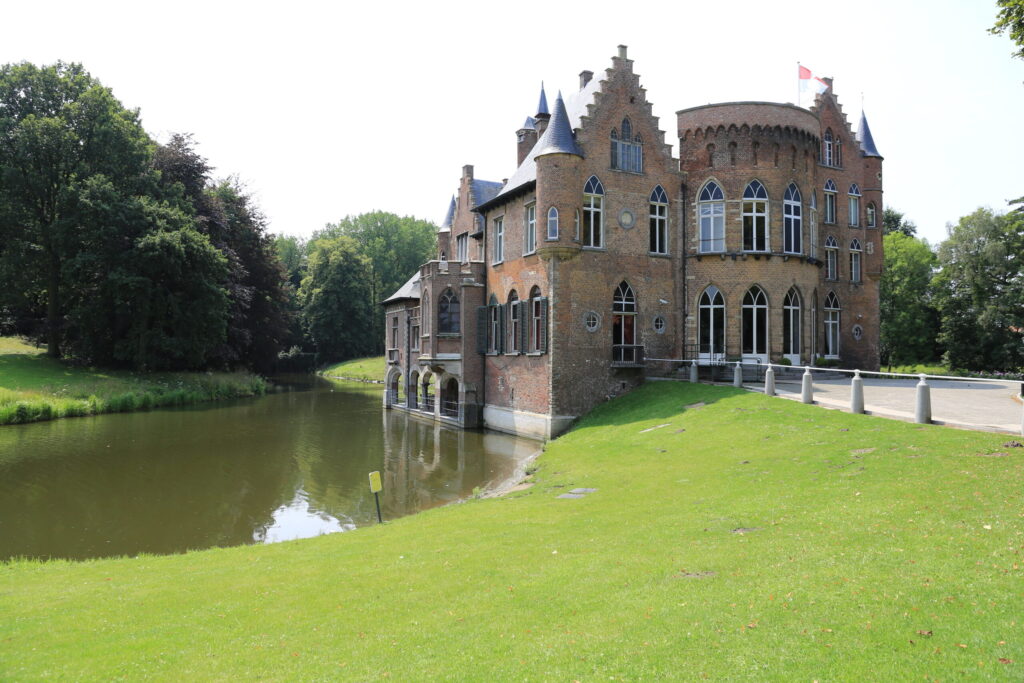 Un château en briques, avec ses tours pointues et ses fenêtres cintrées, se dresse au bord d'un fossé calme et miroitant. De l'herbe verte et des arbres entourent l'édifice sous un ciel dégagé. - Ekilib