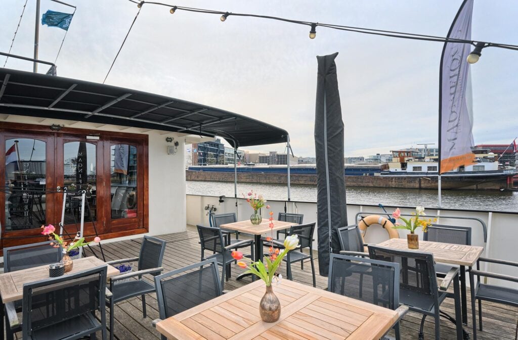 Terrasse extérieure sur un bateau, avec des tables en bois et des chaises en métal ; chaque table est ornée d'un vase rempli de fleurs. La terrasse donne sur une rivière, avec des bâtiments et des bateaux visibles à l'arrière-plan, sous un ciel nuageux. - Ekilib